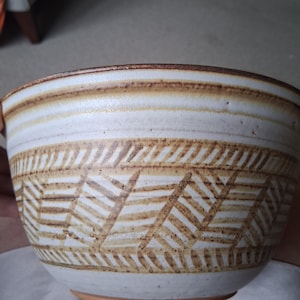 Puede incluir: Un cuenco de cerámica con exterior color crema y un patrón geométrico marrón. El borde y la base del cuenco también son marrones. El cuenco tiene un aspecto rústico y hecho a mano.