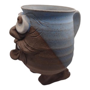 Könnte beinhalten: Eine Keramiktasse mit einem skurrilen Gesicht, mit brauner und blauer Glasur. Die Tasse hat einen Henkel und einen breiten, runden Körper.