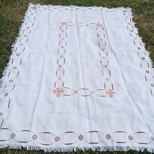 May include: White tablecloth with a brown and orange embroidered floral design. The tablecloth has a fringed edge.