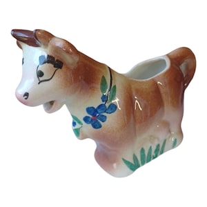 May include: A brown and white ceramic creamer in the shape of a cow with blue flowers and green leaves painted on it.