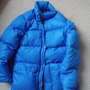 Könnte beinhalten: Eine leuchtend blaue Pufferjacke mit hohem Kragen und zwei Vordertaschen. Die Jacke hat ein gestepptes Design und einen Knopf- und Reißverschluss. Die Jacke besteht aus einem glänzenden, wasserabweisenden Material.