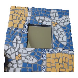 Puede incluir: Espejo cuadrado de mosaico con un espejo central. El mosaico está compuesto por azulejos azules, blancos y amarillos, algunos con diseños florales. Los azulejos están colocados en una lechada gris.