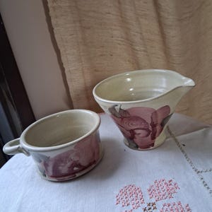 Puede incluir: Un juego de jarra de crema y taza de cerámica. Las piezas de color crema presentan un diseño floral en tonos burdeos y grises. La taza tiene un asa y la jarra de crema tiene un pico. El juego está sobre un mantel blanco.