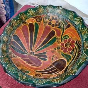 May include: A round ceramic plate with a green, brown, and orange floral design. The plate features a stylized bird with a fan-shaped tail.