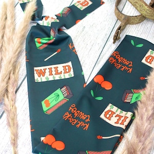 May include: Dark teal scarf with a repeating pattern of matchboxes, cherries, and the text "WILD" and "Kick Rocks Cowboy". The scarf also features a spur and dried ornamental grass.