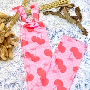 May include: Pink leggings with a red cherry pattern. The leggings have a checkered background and a tie at the top. The fabric is soft and lightweight. The leggings are displayed on a fluffy, white and blue surface.