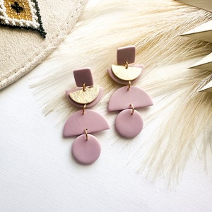 Puede incluir: Pendientes geom&eacute;tricos de color malva con detalles dorados. Cada pendiente presenta un cuadrado, un semic&iacute;rculo y un c&iacute;rculo, unidos por anillas doradas. Se exhiben sobre una superficie blanca con un fondo neutro.