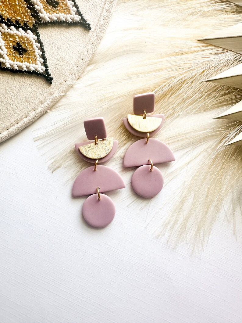 Puede incluir: Pendientes geom&eacute;tricos de color malva con detalles dorados. Cada pendiente presenta un cuadrado, un semic&iacute;rculo y un c&iacute;rculo, unidos por anillas doradas. Se exhiben sobre una superficie blanca con un fondo neutro.