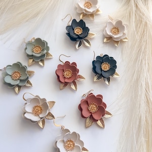 May include: A collection of handmade floral earrings in various colors, including white, sage green, dusty rose, and navy blue. Each earring features a flower design with gold-colored accents and leaf details. The earrings are displayed on a white surface.