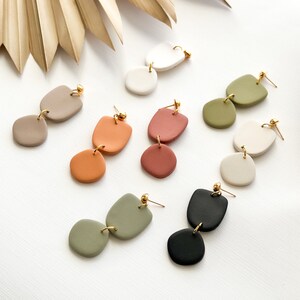 May include: A collection of handmade dangle earrings in various colors, including white, tan, orange, red, green, and black. Each earring features two or three connected, organic-shaped pieces with gold-colored metal accents. The earrings are displayed on a white surface.