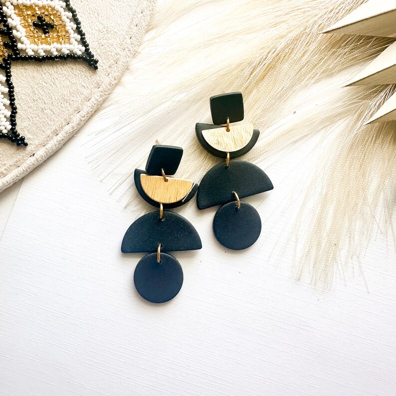 Puede incluir: Un par de pendientes geom&eacute;tricos modernos. Cada pendiente presenta una combinaci&oacute;n de elementos negros y dorados, incluyendo un cuadrado, un semic&iacute;rculo y un c&iacute;rculo, conectados por anillas doradas. Los pendientes est&aacute;n sobre un fondo blanco con detalles de plumas.