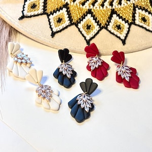 May include: A collection of handmade earrings in various colors, including white, black, and burgundy. Each earring features a fan-shaped design with a cluster of clear crystals. The earrings are displayed on a white surface, with a decorative hat in the background.