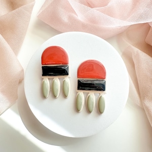 May include: A pair of dangle earrings featuring a glossy red semi-circle top, a horizontal black rectangular middle section, and three pale sage green oval-shaped drops hanging from the bottom, all arranged in a symmetrical geometric design.