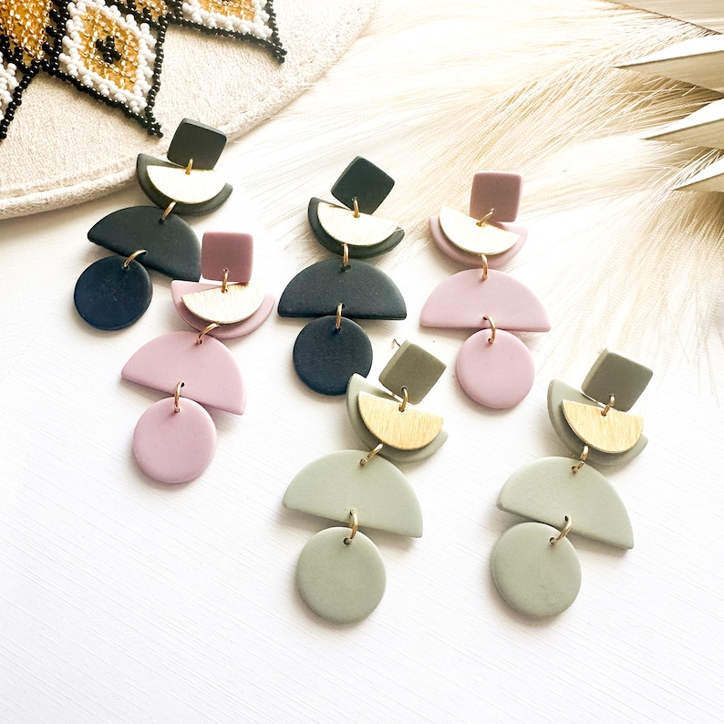 Puede incluir: Pendientes colgantes geom&eacute;tricos en varios colores, incluyendo rosa empolvado, verde salvia y azul marino. Cada pendiente presenta una combinaci&oacute;n de formas: c&iacute;rculos, semic&iacute;rculos y cuadrados, con detalles dorados. Sobre una superficie blanca.