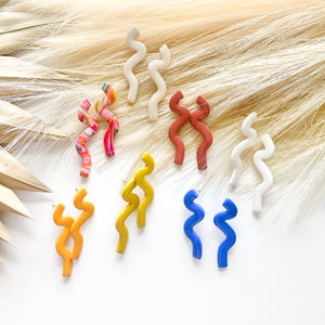 May include: A collection of handmade, abstract stud earrings in various colors. The earrings feature a wavy, elongated design in shades of white, orange, yellow, blue, and a marbled pattern. The earrings are displayed on a white surface with a neutral background.