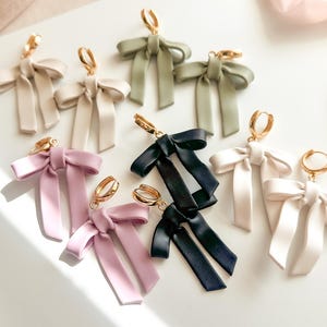 May include: A collection of bow-shaped earrings in various colors including sage green, soft pink, black, and cream. Each earring features a smooth, matte-finish bow suspended from a small, circular gold-toned metal hoop clasp for secure attachment.