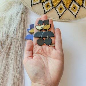 Puede incluir: Pendientes colgantes geom&eacute;tricos en negro, dorado y azul. Los pendientes presentan una combinaci&oacute;n de formas, incluyendo cuadrados, semic&iacute;rculos y c&iacute;rculos. Los pendientes se sostienen en una mano, con un sombrero y plumas en el fondo.