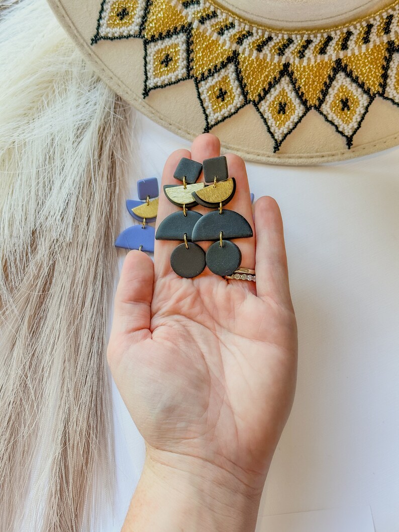 Puede incluir: Pendientes colgantes geom&eacute;tricos en negro, dorado y azul. Los pendientes presentan una combinaci&oacute;n de formas, incluyendo cuadrados, semic&iacute;rculos y c&iacute;rculos. Los pendientes se sostienen en una mano, con un sombrero y plumas en el fondo.