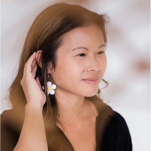 May include: A woman wearing daisy-shaped earrings. The earrings are white with yellow centers and gold hoops. The woman is wearing a black top and has long brown hair. The background is blurred and light.