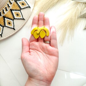 May include: A pair of bright yellow, abstract-shaped earrings with gold-colored hooks. The earrings are held in a hand, with a white and gold-colored hat in the background. The earrings have a modern, artistic design.