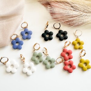 May include: A collection of flower-shaped earrings in various colors, including blue, black, white, green, red, and yellow. Each earring features a gold-colored hoop. The earrings are displayed on a white surface.