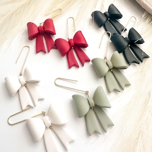 May include: A collection of bow-shaped earrings in various colors: red, black, white, and sage green. Each earring features a gold-colored hook. The bows have a matte finish and are arranged on a white surface.
