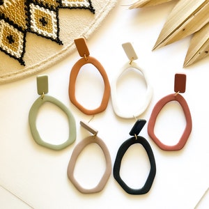 May include: A collection of six pairs of abstract, oval-shaped earrings in various colors, including green, tan, white, black, and shades of brown. Each earring has a rectangular top and a gold-colored post. The earrings are displayed on a white surface.