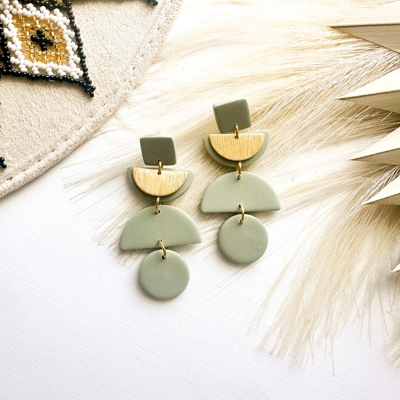 Puede incluir: Un par de pendientes colgantes con un dise&ntilde;o moderno. Cada pendiente presenta un cuadrado, un semic&iacute;rculo dorado, un semic&iacute;rculo verde salvia y un c&iacute;rculo verde salvia, todos conectados por aros dorados. Fondo blanco.