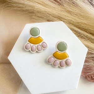 May include: A pair of handmade stud earrings. Each earring features a light green circle, a yellow half-circle, and a row of pink, rounded shapes. The earrings are set on a white, geometric display.