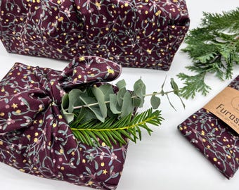 May include: Burgundy fabric gift wraps with a floral pattern, including a wrapped package with greenery and a small folded wrap. The fabric features a design of white, yellow, and pink floral elements. A small package with the text "Furoshiki" is also visible.