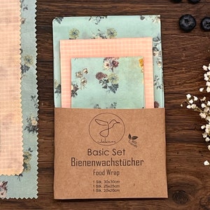 Peut inclure: Un ensemble de trois emballages alimentaires en cire d'abeille à motifs floraux. Les emballages sont de différentes tailles : 30 x 30 cm, 25 x 25 cm et 20 x 20 cm. Les emballages sont emballés dans un sac en papier brun avec le nom de la marque "Jakan" et le texte "Basic Set Bienenwachstücher Food Wrap" imprimé dessus.