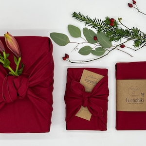 Könnte beinhalten: Drei rote Stoffgeschenkverpackungen, eine mit einem Blütenstiel und zwei mit einer Schleife. Die Verpackungen sind auf einer weißen Oberfläche mit Zweigen und Beeren angeordnet. Die Verpackungen sind mit dem Wort "Furoshiki" beschriftet.