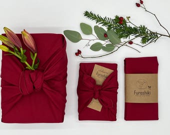 Puede incluir: Tres envoltorios de regalo de tela roja, uno con un tallo de flor y dos con un lazo. Los envoltorios están dispuestos sobre una superficie blanca con ramitas de vegetación y bayas. Los envoltorios están etiquetados con la palabra "Furoshiki".