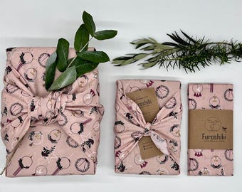 May include: Three pink Furoshiki fabric gift wraps with a repeating pattern of wreaths, stars, and botanical elements. The wraps are tied in various ways, with sprigs of greenery adding a natural touch. One wrap has a brown tag.
