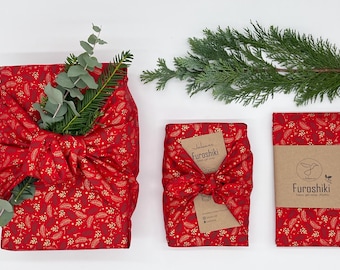 May include: Red fabric gift wraps with gold floral patterns, tied with bows. One wrap is decorated with greenery. A smaller wrap has a tag that says "Furoshiki". The wraps are on a white surface.