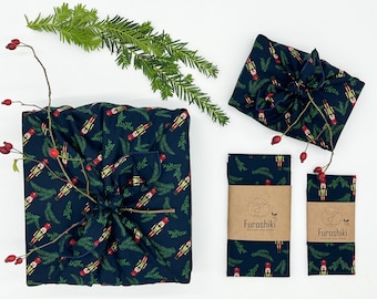 Puede incluir: Tres paquetes de envoltura de regalo de tela azul con un patrón de cascanueces y ramas de pino. El paquete más grande está atado con una cinta roja. Los paquetes más pequeños tienen una etiqueta marrón con el texto "Furashiki" y un logotipo de hoja verde.