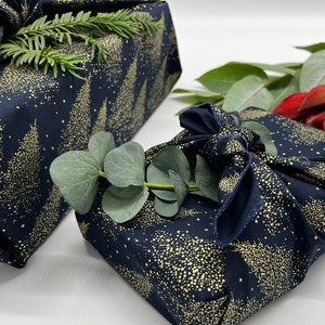 May include: Two wrapped gifts with navy blue fabric featuring a gold tree and dot pattern. One is decorated with a sprig of greenery, the other with eucalyptus leaves and red flowers. The gifts are presented on a white surface.