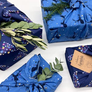 Könnte beinhalten: Vier Geschenke, verpackt in blauem und marineblauem Stoff mit einem Weihnachtsbaum- und Rentiermuster. Der Stoff ist mit einem Knoten gebunden und mit grünen Zweigen verziert. Ein Geschenk hat ein braunes Etikett mit dem Text "Furoshiki Japanese Fabric Gift Wrap".