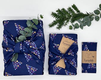 Puede incluir: Tres regalos envueltos en tela azul con un patrón de árbol de Navidad. La tela está atada con un nudo y decorada con una pequeña etiqueta marrón que dice "Furoshiki japonés". Los regalos están sobre una superficie blanca con ramas verdes.