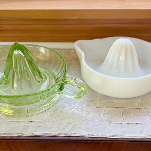 May include: Two citrus juicers, one made of translucent light green glass and the other made of opaque white milk glass. Each juicer features a central reamer, a circular basin with ribbed sides, a small pouring spout, and a side handle for gripping.