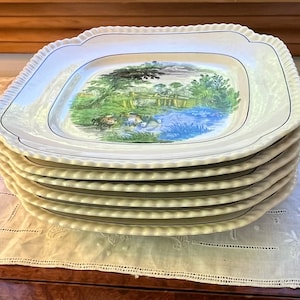 May include: A stack of six white square plates with a scalloped edge and a landscape scene in the center. The plates have a blue border and a detailed illustration of a pond with trees and ducks.
