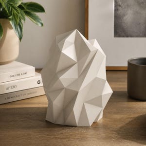 May include: A geometric, off-white sculpture with a faceted surface, resembling a stylized mountain or rock formation. The sculpture is placed on a wooden surface, with books and a plant visible in the background. The sculpture is a decorative home accent.
