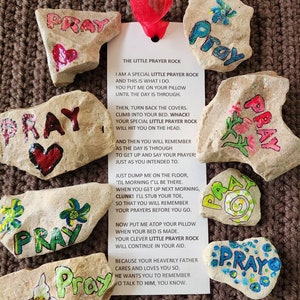 Puede incluir: Un conjunto de piedras pintadas con la palabra "Rezar" en cada una. Las piedras est&aacute;n decoradas con flores, corazones y espirales. Se incluye una tarjeta con instrucciones para usar las piedras como recordatorios de oraci&oacute;n. La tarjeta dice "La peque&ntilde;a piedra de oraci&oacute;n" e incluye un poema sobre el uso de las piedras para recordar rezar.