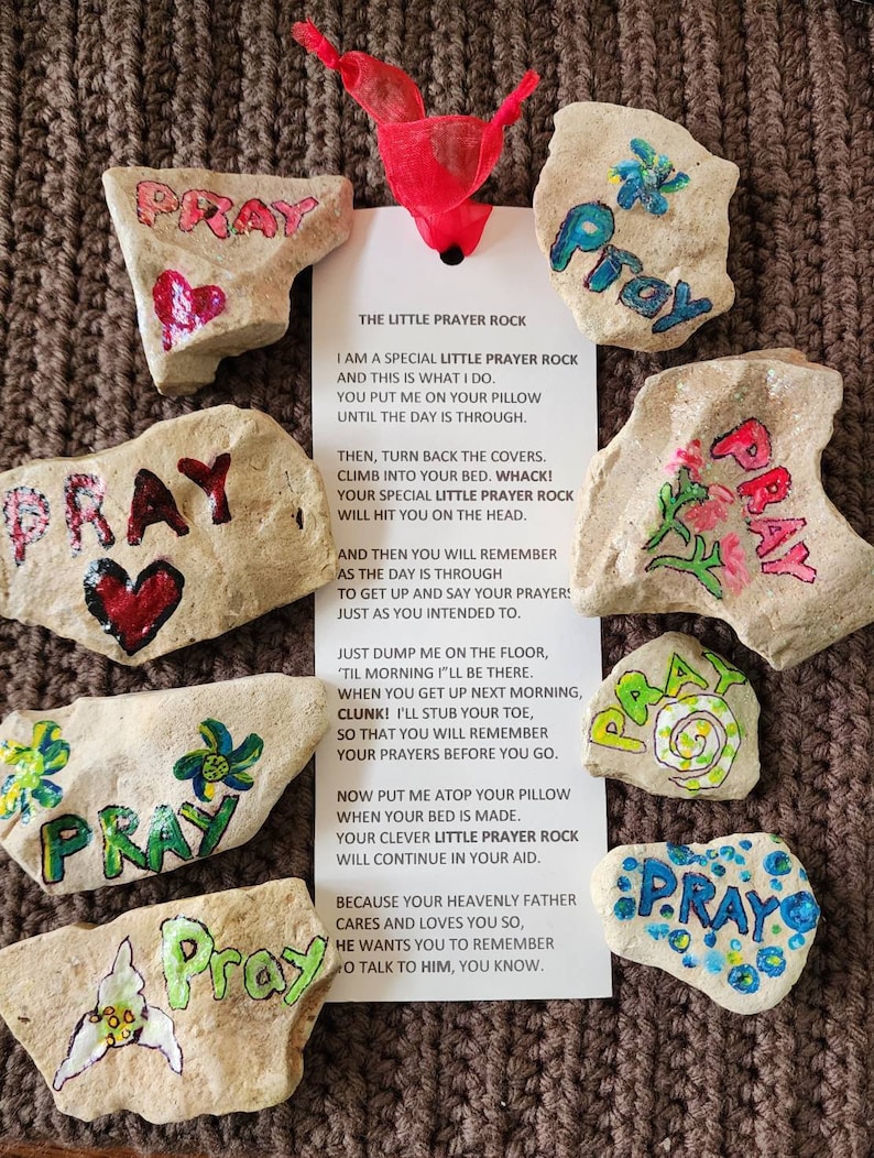 Puede incluir: Un conjunto de piedras pintadas con la palabra "Rezar" en cada una. Las piedras est&aacute;n decoradas con flores, corazones y espirales. Se incluye una tarjeta con instrucciones para usar las piedras como recordatorios de oraci&oacute;n. La tarjeta dice "La peque&ntilde;a piedra de oraci&oacute;n" e incluye un poema sobre el uso de las piedras para recordar rezar.
