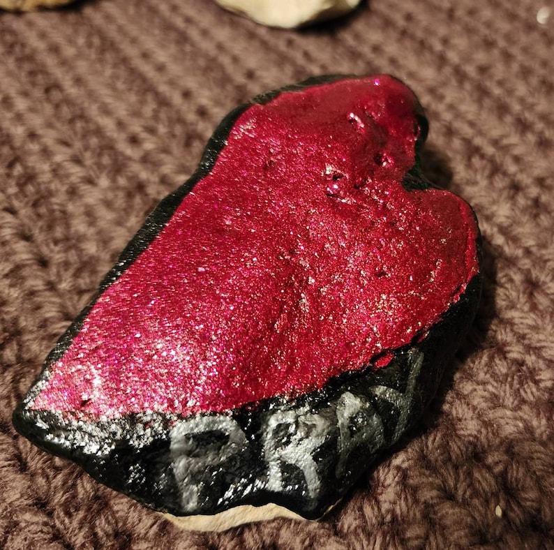 Puede incluir: Una piedra en forma de coraz&oacute;n pintada de rojo con purpurina y pintura negra con la palabra "Frank" escrita en negro.