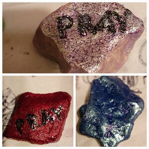 Puede incluir: Tres piedras pintadas con purpurina. La primera piedra es morada con la palabra "PMA" escrita en negro. La segunda piedra es roja con la palabra "PRETTY" escrita en negro. La tercera piedra es azul con un acabado brillante.
