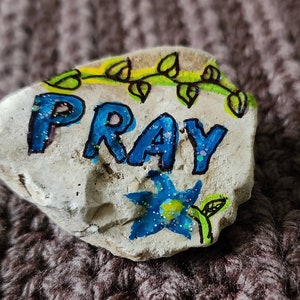 Puede incluir: Una roca blanca pintada con la palabra "PRAY" en azul con detalles de purpurina. Una vid verde con hojas est&aacute; pintada sobre la palabra y una flor azul con centro amarillo est&aacute; pintada debajo de la palabra.