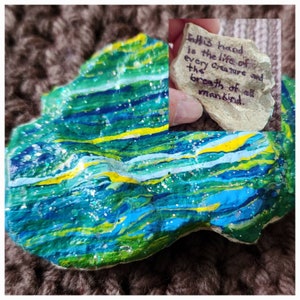 Puede incluir: Una piedra pintada con rayas azules, verdes, amarillas y blancas. La piedra tiene brillantina incrustada en la pintura. La piedra tiene un mensaje escrito en ella: "In this hand is the life of every creature and the breath of all mankind."