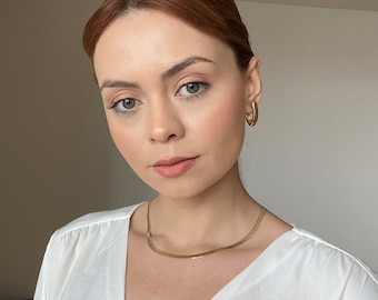 Op de afbeelding: Een vrouw met rood haar draagt een wit shirt en een gouden kettingketting. Ze draagt gouden oorringen.