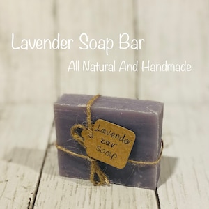 Pu&ograve; includere: Una saponetta di colore lavanda avvolta in spago con un'etichetta di carta marrone su cui &egrave; scritto "Lavender bar soap". La saponetta &egrave; su una superficie di legno bianco. Il testo "Lavender Soap Bar" e "All Natural And Handmade" &egrave; scritto in bianco su uno sfondo bianco sopra la saponetta.